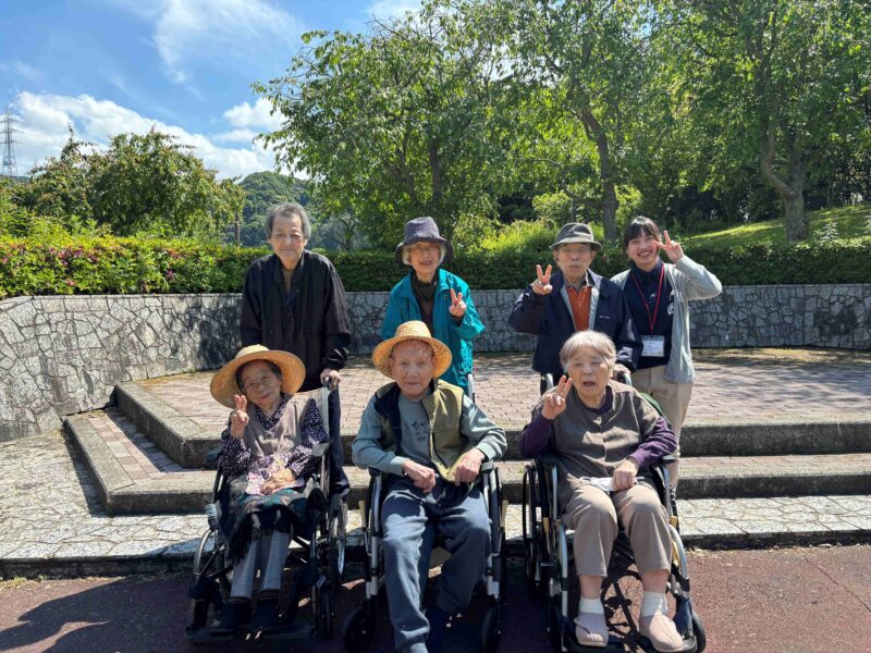 介護施設の入居者と職員が屋外でレクリエーション活動を楽しむ風景。車いす利用者も含め、皆笑顔で記念撮影。